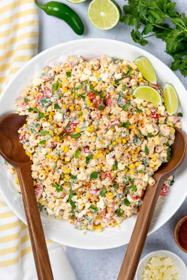 Large serving bowl of Mexican street corn pasta salad garnished with chili powder and surrounded by fresh cilantro, lime wedges, jalapeños, cotija cheese, and a bowl of chili powder.