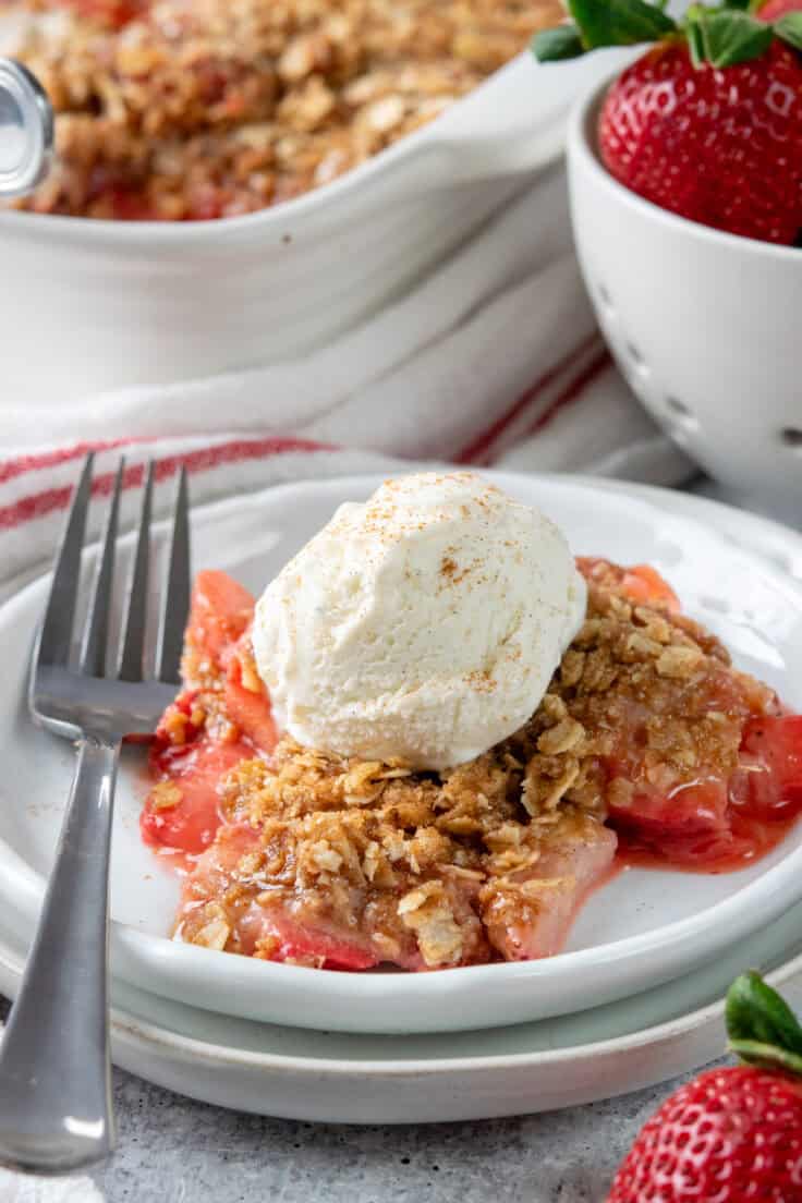 Large serving of homemade strawberry crisp with vanilla ice cream on a dessert plate beside the baking dish and a bowl filled with fresh ripe strawberries.