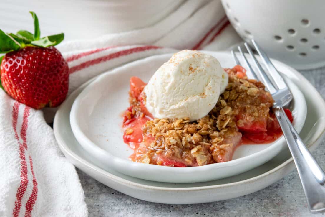 Dessert plate with a generous serving of strawberry crisp topped with vanilla ice cream beside a fork and a ripe fresh strawberry.
