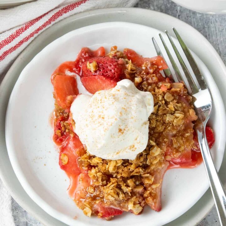 Overhead view of homemade strawberry crisp with a buttery oat crumble topping and melting vanilla ice cream served beside the baking dish filled with the warm summer dessert.