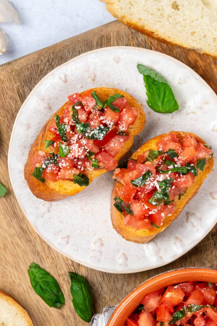 Tomato bruschetta appetizer served on a plate beside fresh basil leaves and a bowl of tomato, basil, and garlic topping.