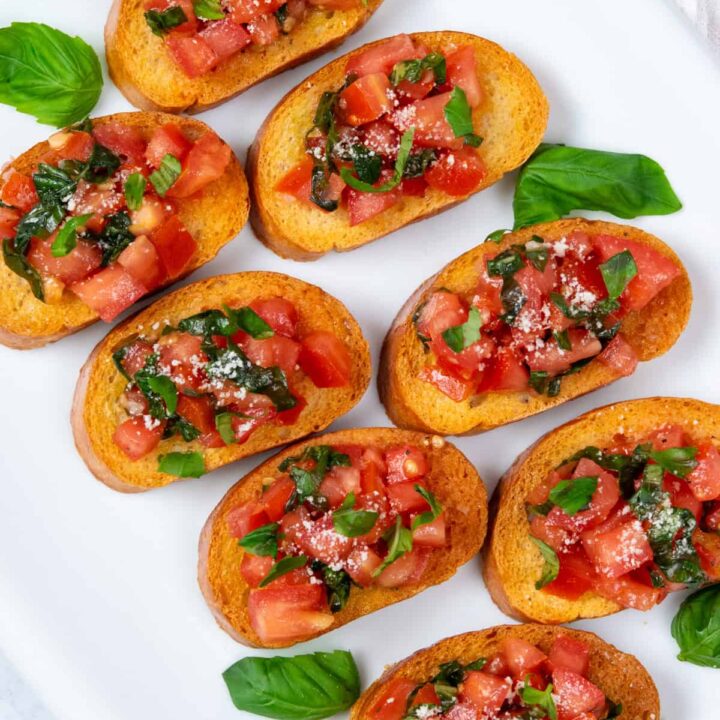 Classic Italian tomato bruschetta piled onto toasted bread on a white platter with fresh basil leaves and grated parmesan cheese on top.