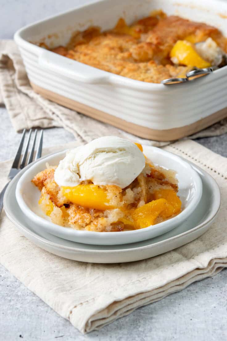 A serving of Bisquick peach cobbler with vanilla ice cream on a dessert plate beside the baking dish, showing juicy peaches and a buttery cinnamon-speckled cobbler.