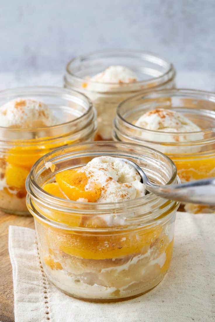 Four mason jars filled with homemade Bisquick peach cobbler, each topped with vanilla ice cream and a juicy peach slice.
