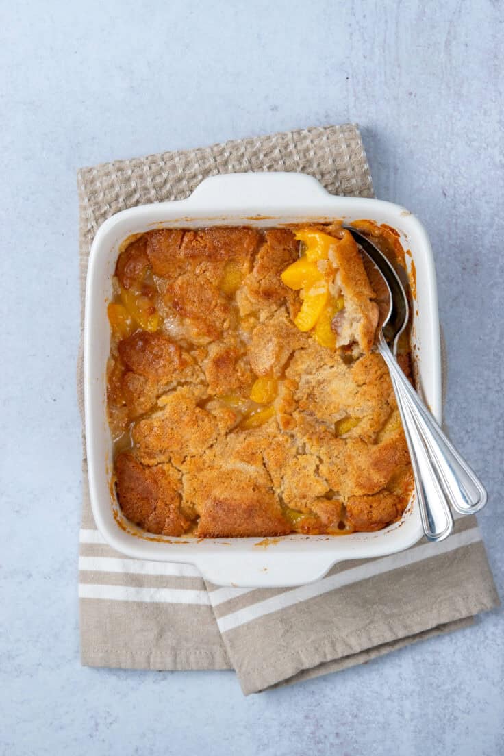 Golden brown Bisquick peach cobbler fresh from the oven with serving spoons lifting out a warm scoop of buttery cobbler and peaches.
