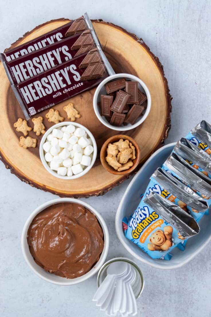 Walking s'mores dessert bar with bowls of marshmallows, chopped Hershey's chocolate, Teddy Grahams, chocolate pudding, prepared snack bags, and spoons for building individual desserts.