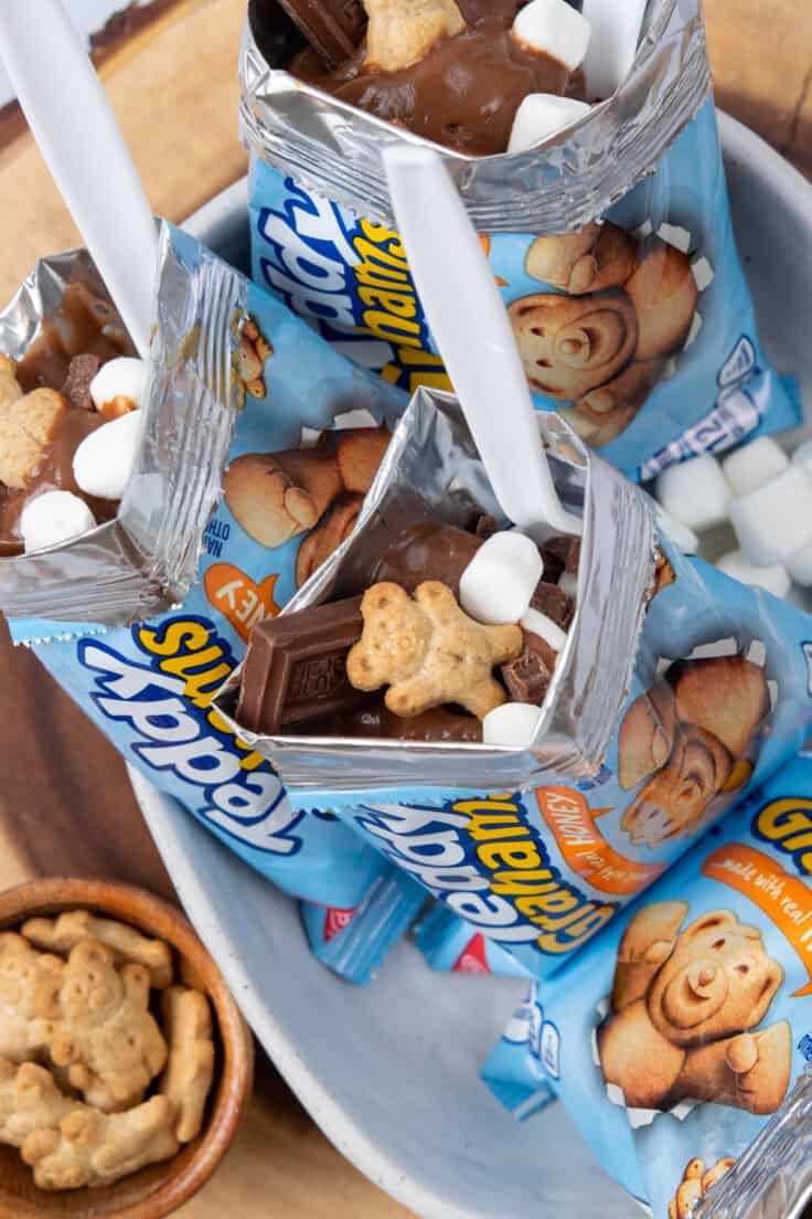 Five assembled walking s'mores bags garnished with Teddy Grahams and Hershey's chocolate, displayed on a serving platter with marshmallows and extra toppings nearby.