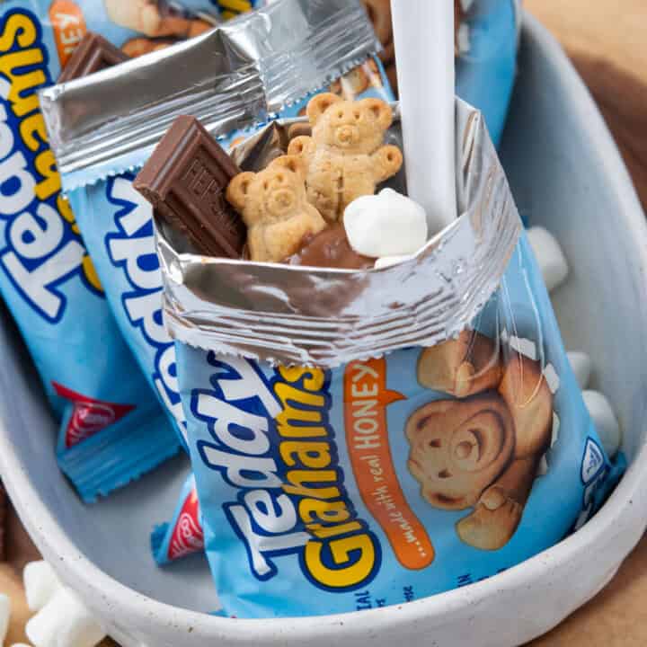 Walking s'mores in Teddy Grahams snack bags topped with chocolate pudding, mini marshmallows, Hershey's chocolate, and Teddy Grahams, each served with a spoon.