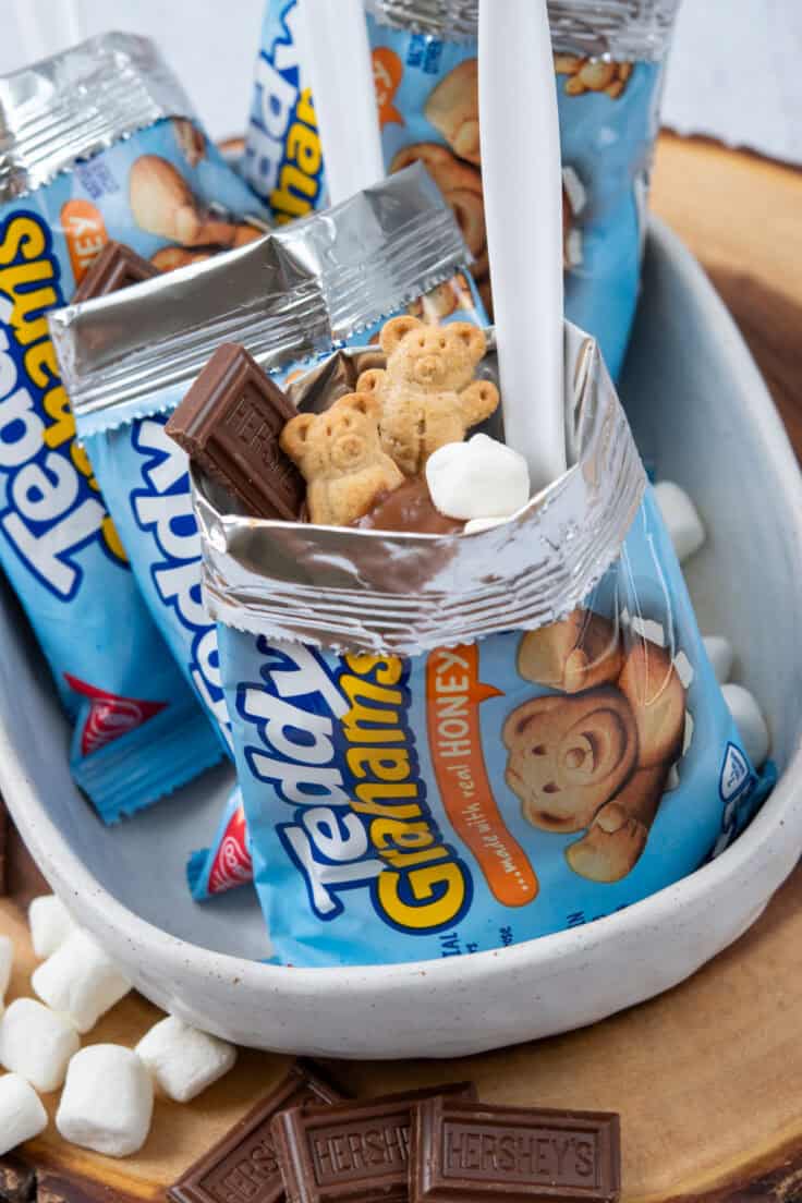 Walking s'mores in Teddy Grahams snack bags topped with chocolate pudding, mini marshmallows, Hershey's chocolate, and Teddy Grahams, each served with a spoon.