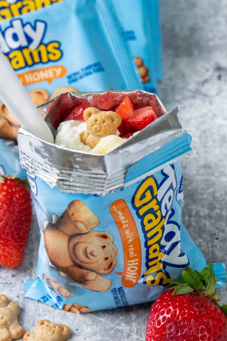 Walking strawberry cheesecake served in a Teddy Grahams snack bag and garnished with whipped cream, strawberries, and cookies, leaning against additional finished dessert bags.