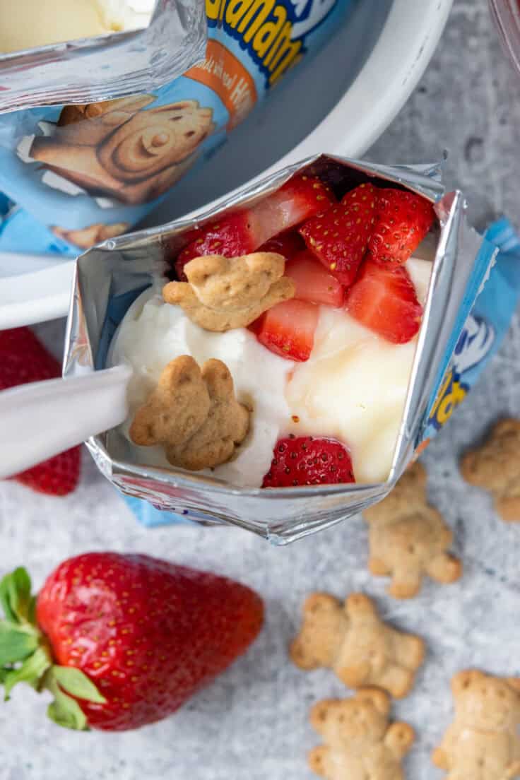 Top-down view of a walking strawberry cheesecake made in a Teddy Grahams bag with cheesecake pudding, juicy strawberries, whipped cream, and Teddy Graham crackers, surrounded by extra cookies and a fresh strawberry.
