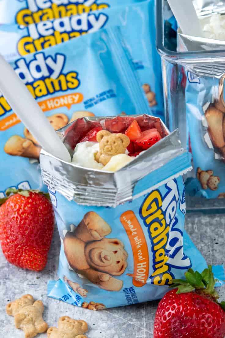 Walking strawberry cheesecake made in a Teddy Grahams bag displayed on a dessert bar alongside the pudding, strawberries, whipped topping, and graham cracker bags used to make it.