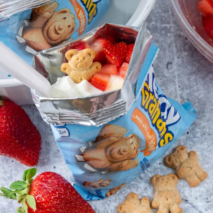 Walking strawberry cheesecake made in a Teddy Grahams snack bag topped with whipped cream, a fresh strawberry slice, a Teddy Graham cracker, and a spoon for serving.