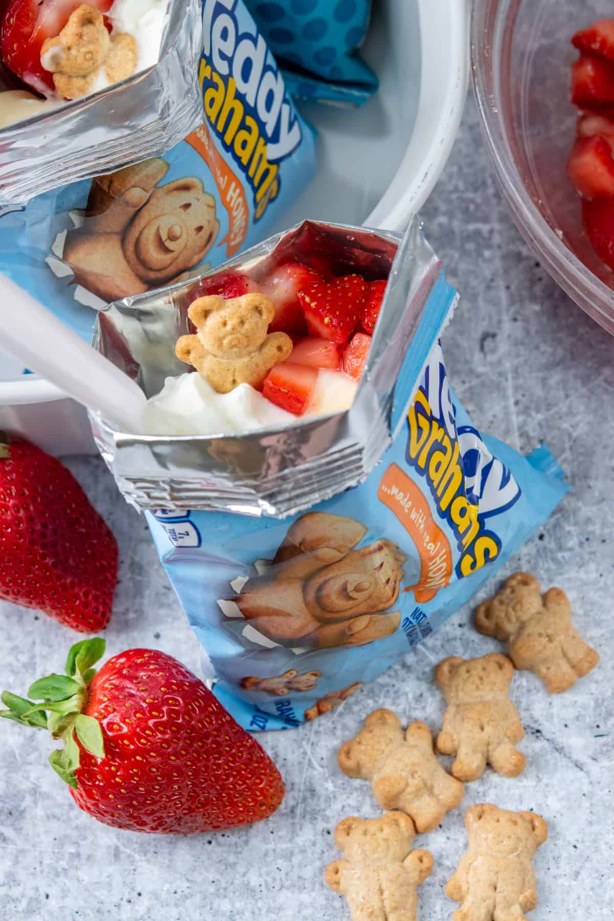Walking strawberry cheesecake made in a Teddy Grahams snack bag topped with whipped cream, a fresh strawberry slice, a Teddy Graham cracker, and a spoon for serving.