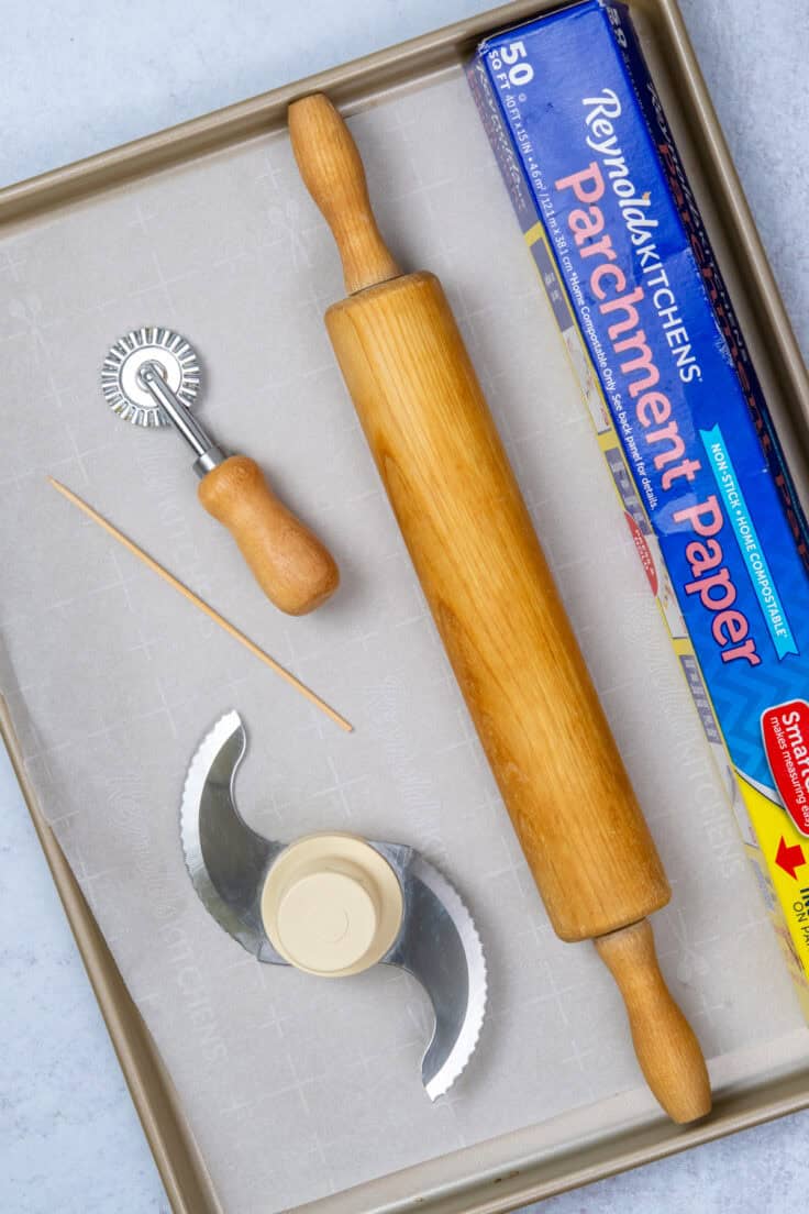 Equipment needed to make homemade Cheez-Its, including a food processor, baking sheet, parchment paper, fluted pastry wheel, and wooden skewer.