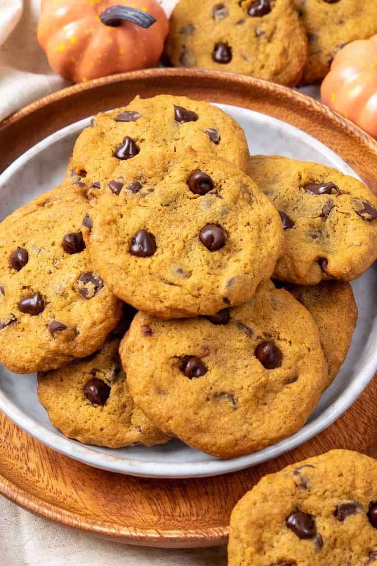 An orange plate of pumpkin chocolate chip cookies surrounded by decorative pumpkins, with additional cookies in the background.