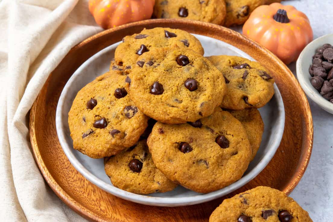 Homemade pumpkin chocolate chip cookies arranged on a serving plate beside a bowl of chocolate chips and decorative pumpkins.