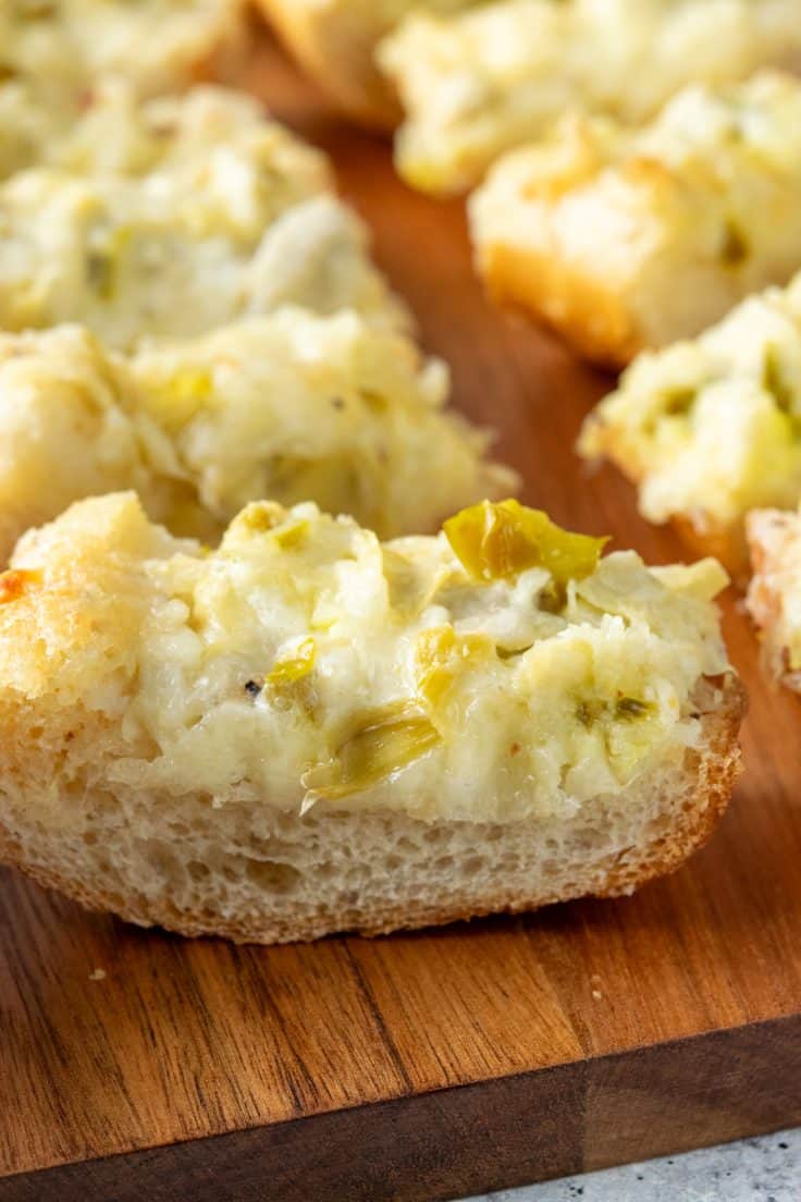 Cheesy artichoke bread served on a wooden board, with a close-up showing melted cheese, artichokes, and bits of jalapeño throughout the appetizer.