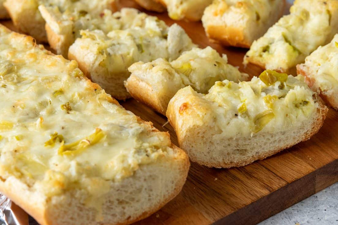 Artichoke bread served on a wooden board, cut into appetizer-size pieces for easy finger-food serving, with creamy artichoke dip baked onto French bread.