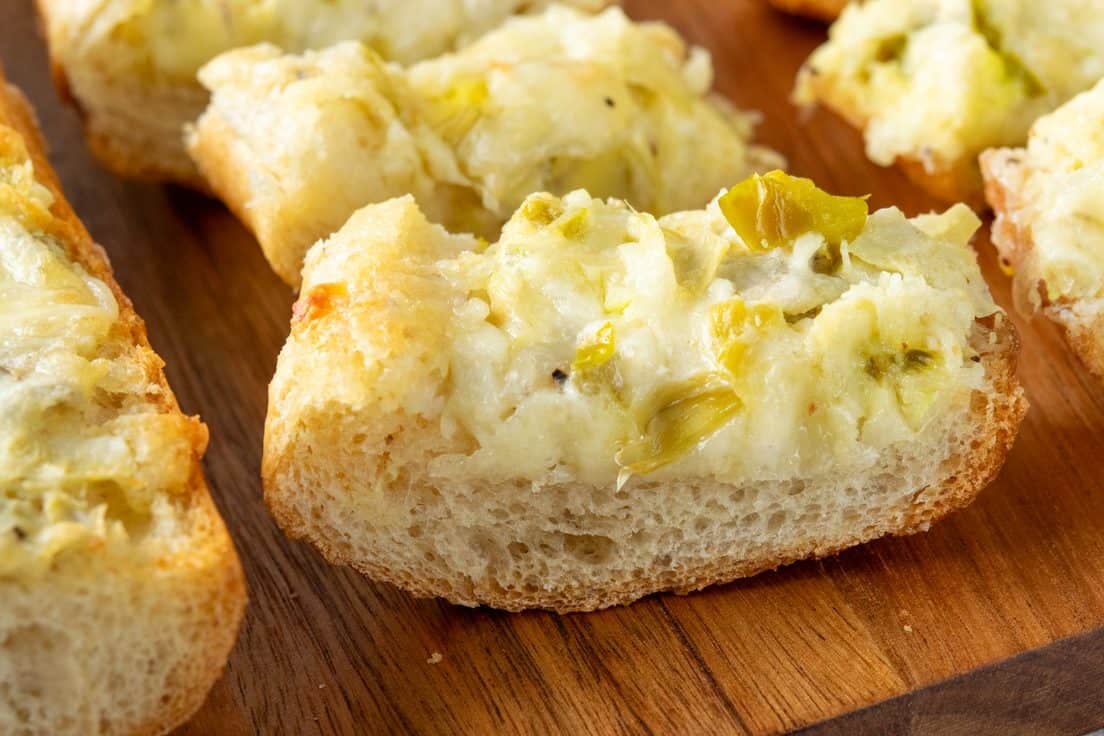 Close-up of a piece of artichoke bread with crisp edges, buttery melted cheese, chopped artichokes, and jalapeños baked into the creamy topping.