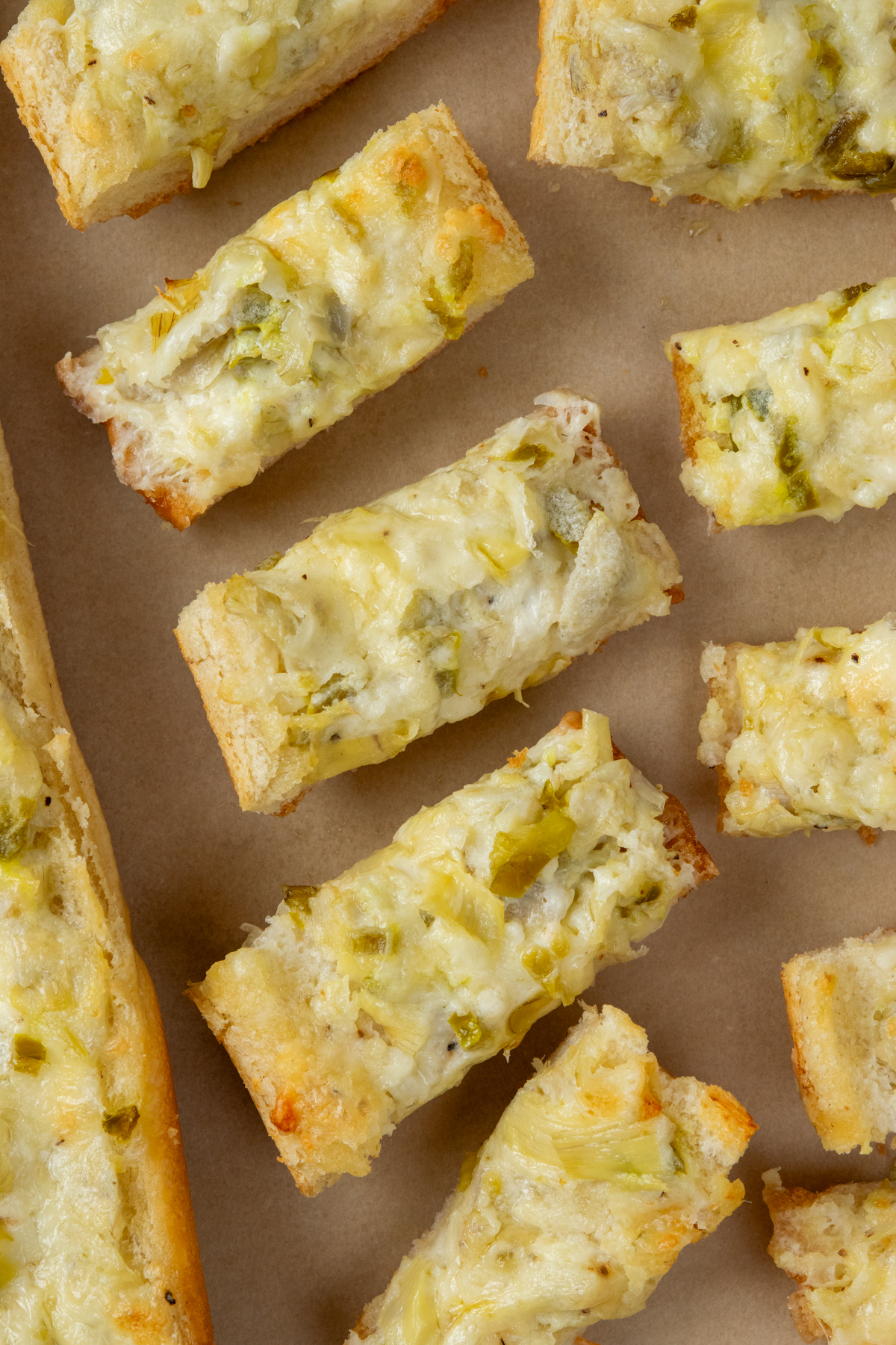 Sliced artichoke bread in appetizer-size pieces alongside a larger piece of French bread topped with melted cheese, chopped artichokes, and jalapeño peppers.