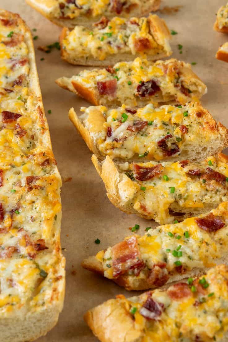 Two loaves of bacon ranch cheese bread on parchment paper, one whole and one sliced into serving pieces, topped with melted cheese, bacon, and fresh chopped chives.