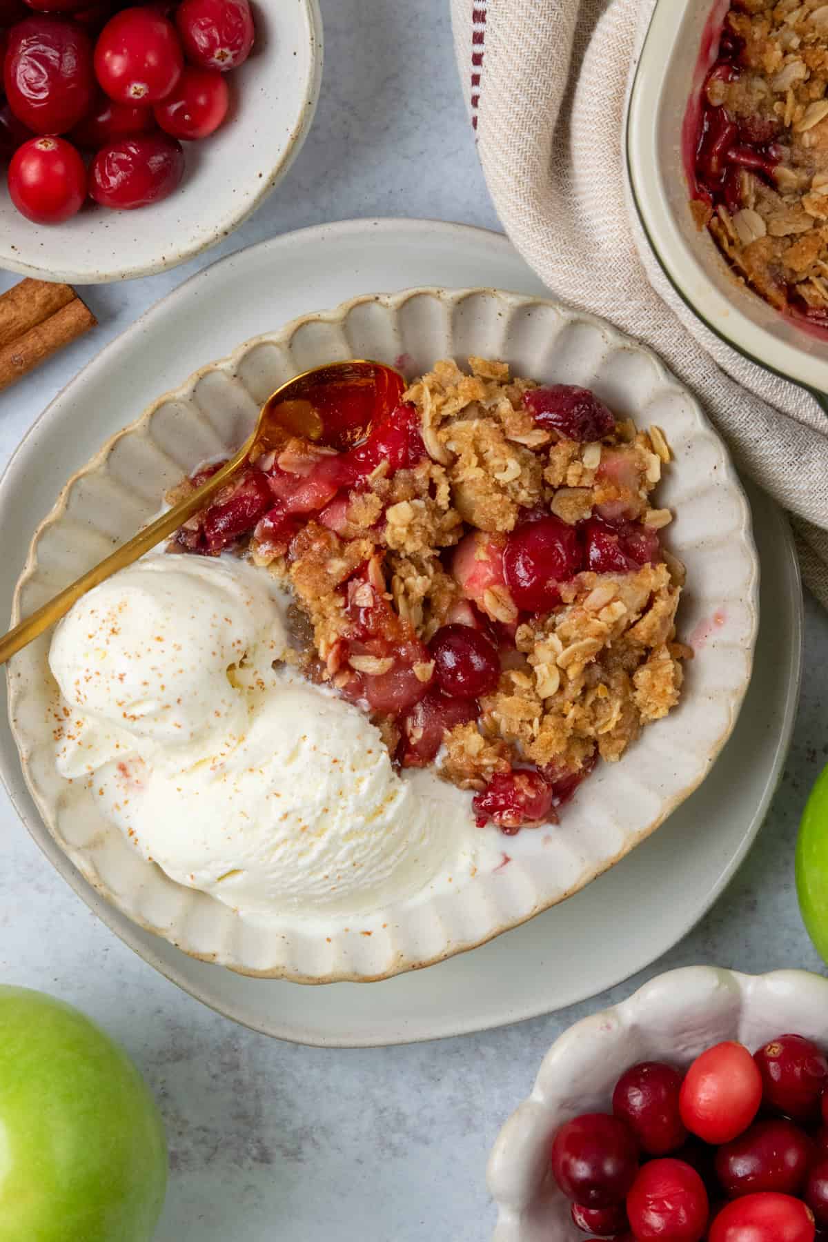 A serving of apple cranberry crisp topped with melting vanilla ice cream, displayed beside a baking dish of the finished dessert, fresh cranberries, and Granny Smith apples.