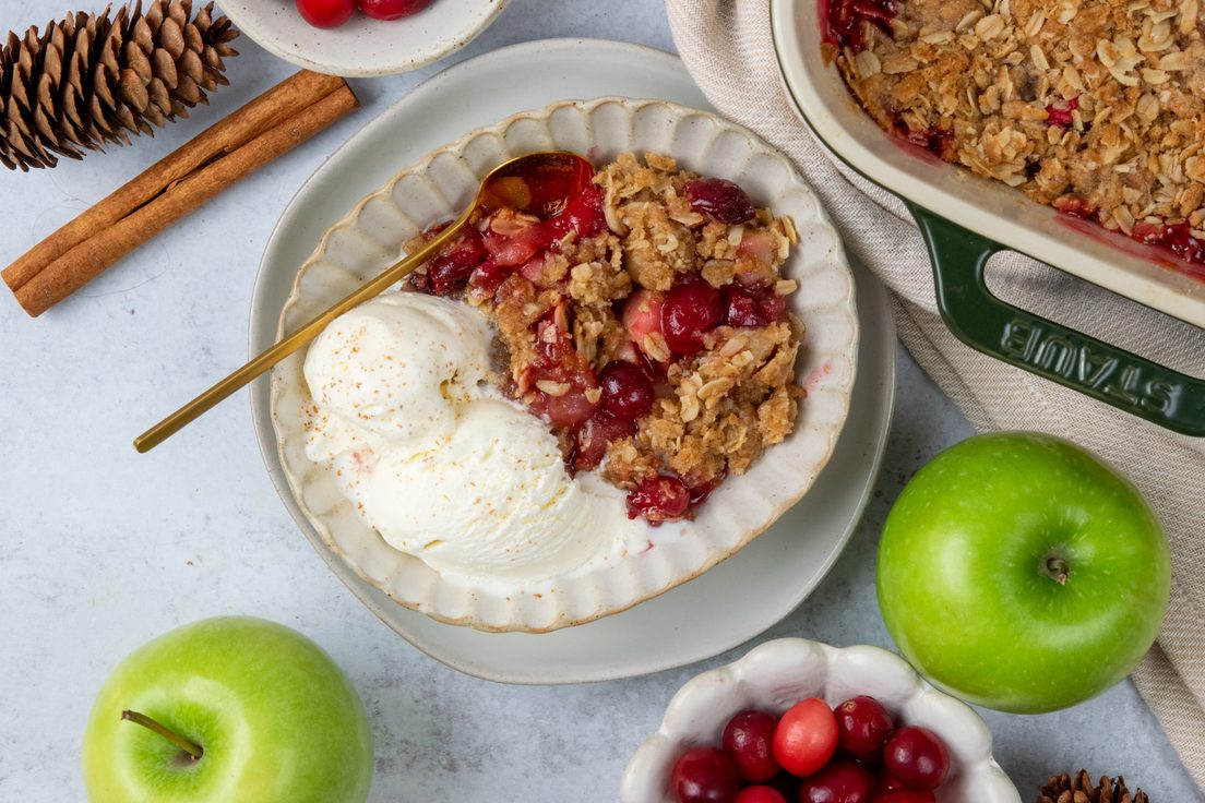 Apple cranberry crisp served warm with vanilla ice cream beside a baking dish of the finished dessert, surrounded by bowls of fresh cranberries, Granny Smith apples, cinnamon sticks, and pinecones for a festive Thanksgiving or Christmas dessert display.