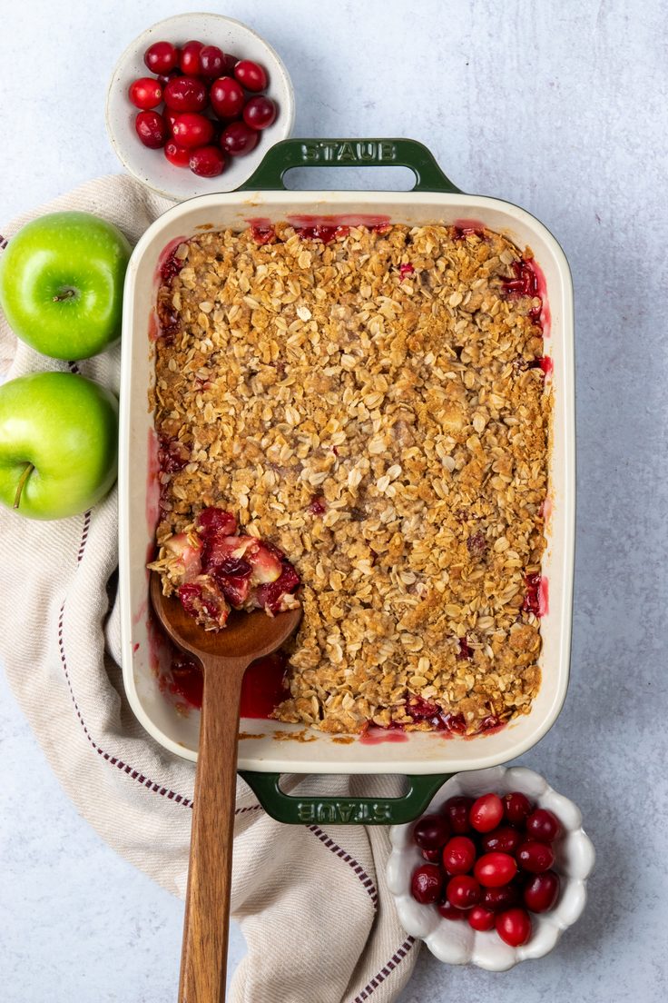 Freshly baked apple cranberry crisp in a baking dish, with red cranberries bubbling around the edges of the golden oat topping and a serving spoon ready to scoop, displayed beside fresh cranberries and Granny Smith apples.