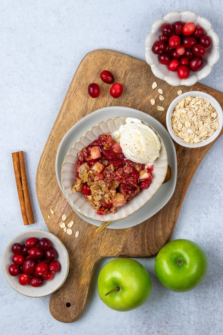 A bowl of apple cranberry crisp topped with vanilla ice cream on a wooden tray, surrounded by fresh cranberries, apples, rolled oats, and cinnamon sticks for a seasonal fall dessert presentation.