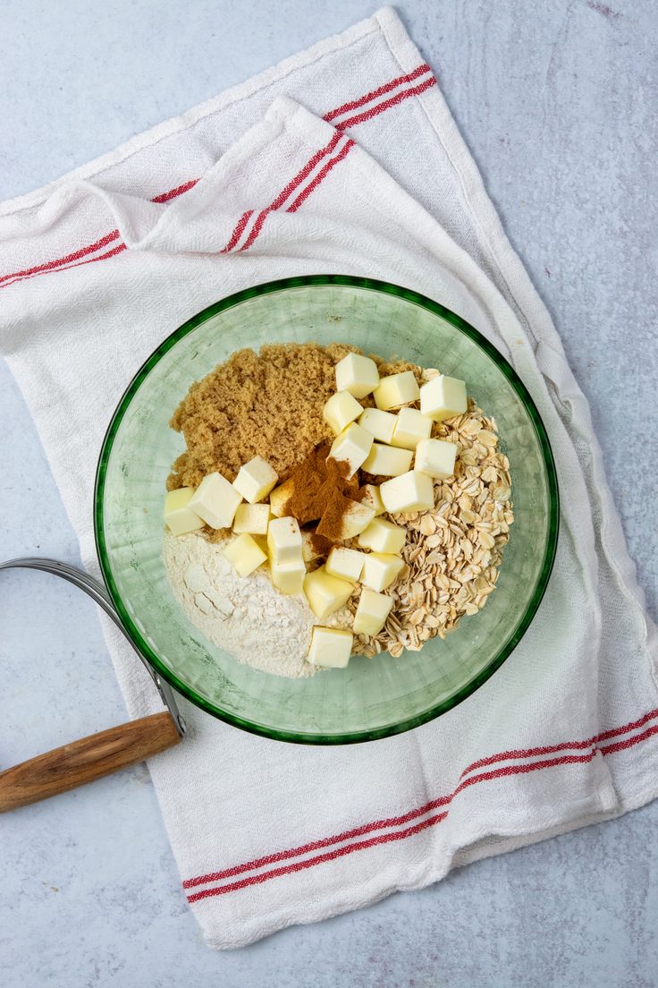 Rolled oats, brown sugar, flour, cinnamon, and small pieces of cold butter in a mixing bowl before the butter has been cut into the dry ingredients.