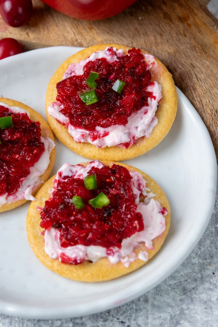 Three buttery crackers on an appetizer plate topped with pink cream cheese and cranberry jalapeno relish, finished with small pieces of diced jalapeno for a sweet and spicy holiday bite.