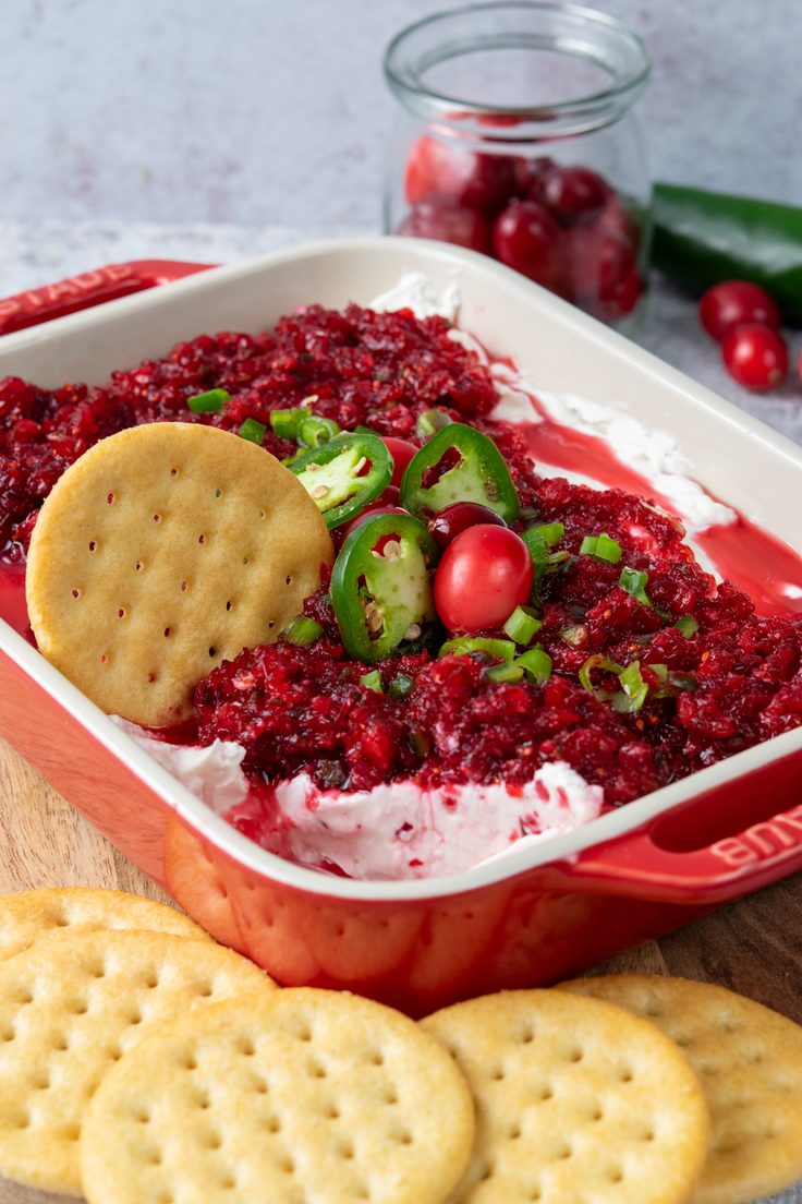Festive cranberry jalapeno dip in a serving dish with a portion scooped out to reveal the creamy cream cheese layer beneath the sweet-tart cranberry topping, surrounded by buttery crackers for dipping.