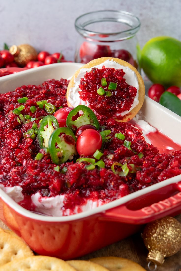 Festive cranberry jalapeno cream cheese dip served in a red dish that's surrounded by crackers for dipping and holiday ornaments.