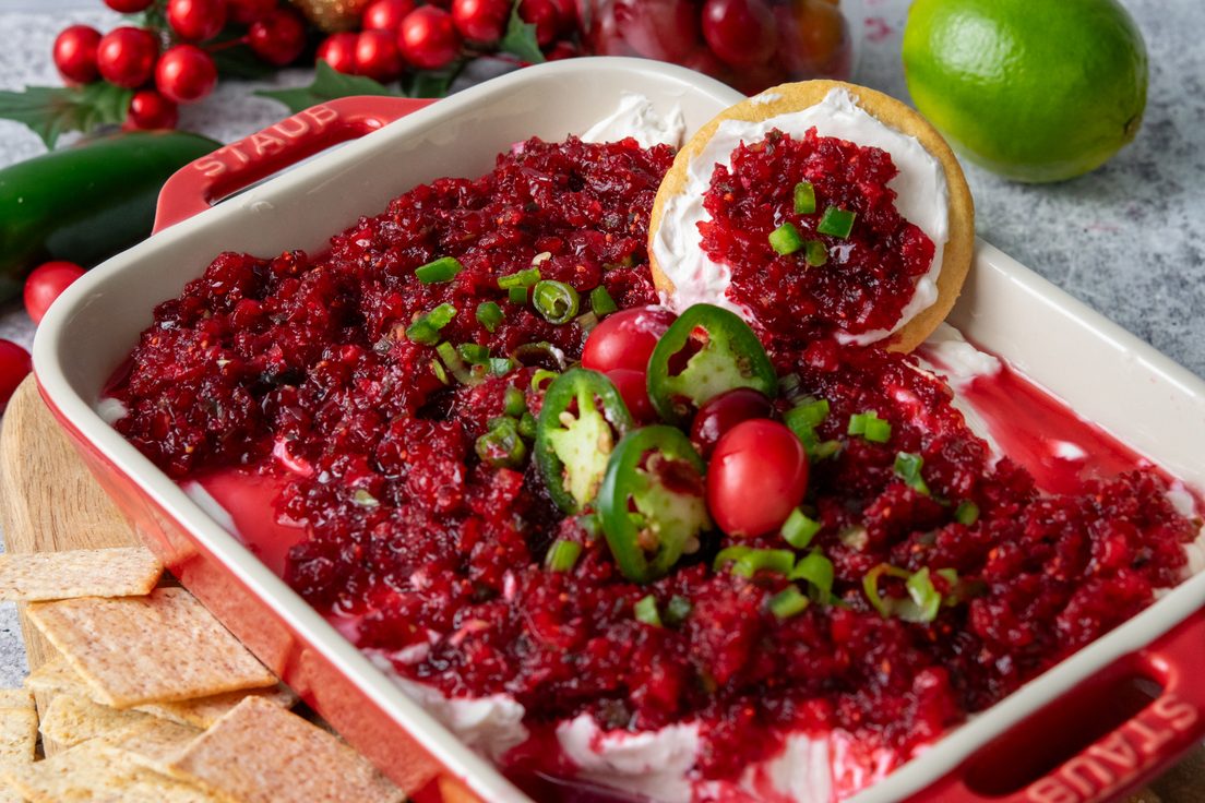 Cranberry jalapeno cream cheese dip in a red serving dish surrounded by crackers, with a deep red cranberry topping layered over cream cheese, garnished with whole cranberries and jalapeno slices, and fresh lime and jalapeno in the background.