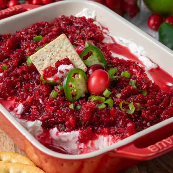 Cranberry jalapeno cream cheese dip layered in a red serving dish, garnished with fresh cranberries and jalapeno slices, with a cracker scooping up the sweet and spicy topping and more crackers arranged around the dish.