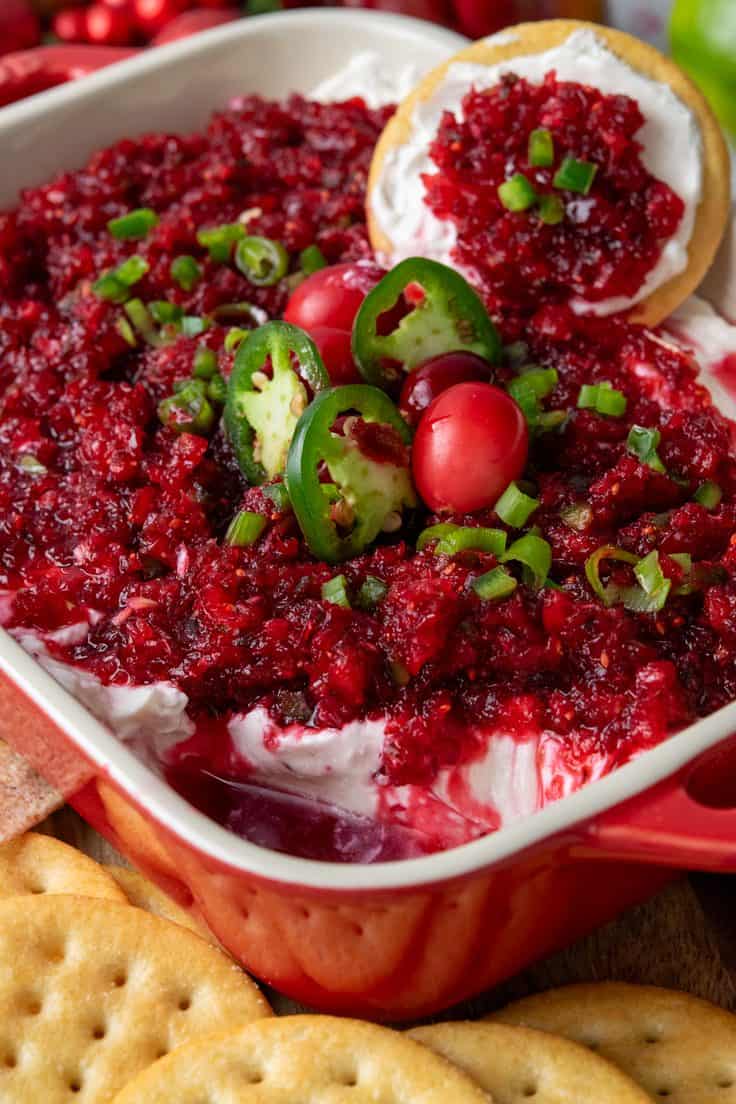 Close-up of cranberry jalapeno cream cheese dip showing chunky red cranberry relish dripping into the creamy cheese layer where a cracker has scooped out a bite, with buttery crackers arranged around the serving dish.