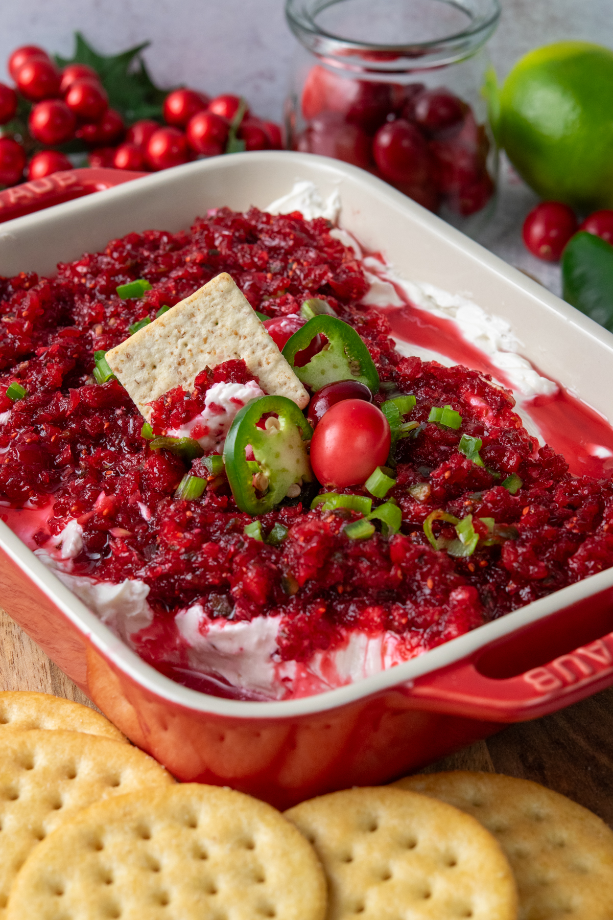 Cranberry jalapeno cream cheese dip layered in a red serving dish, garnished with fresh cranberries and jalapeno slices, with a cracker scooping up the sweet and spicy topping and more crackers arranged around the dish.