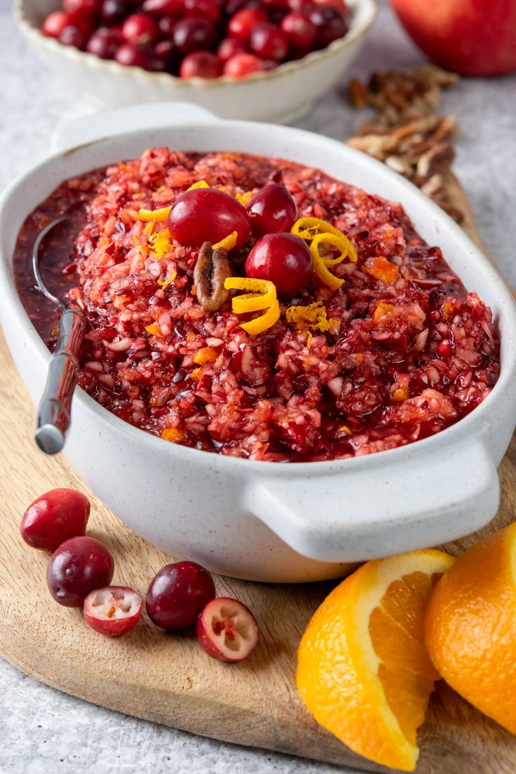 A spoon resting in a bowl of homemade cranberry relish made with fresh cranberries, apple, and orange, surrounded by fresh cranberries, orange slices, and pecans.