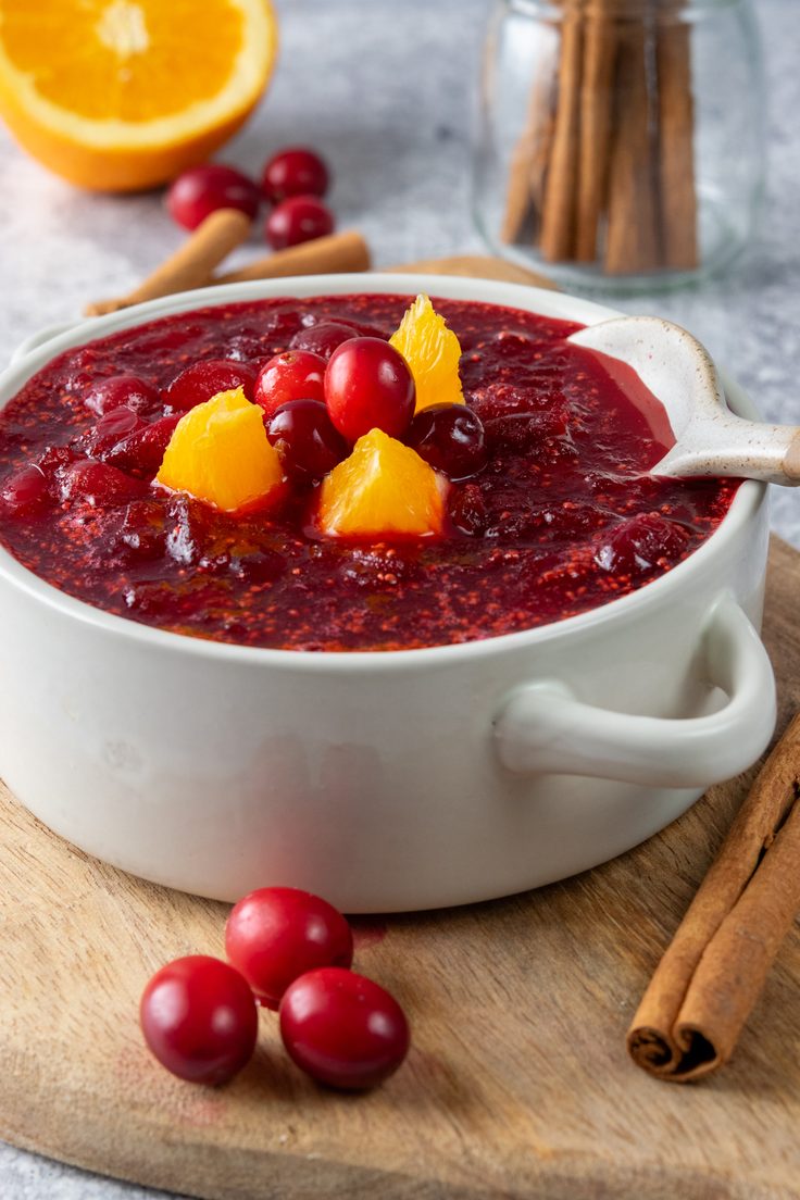 Deep red homemade cranberry orange sauce in a white serving bowl, garnished with orange pieces and whole cranberries, with fresh orange halves and cinnamon sticks arranged behind it for a holiday meal.