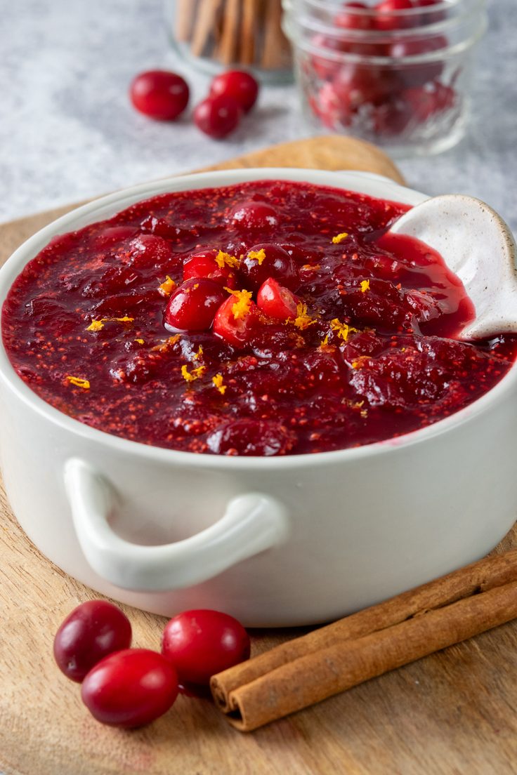 Homemade Thanksgiving cranberry orange sauce garnished with orange zest in a small white bowl with a spoon, surrounded by a jar of cinnamon sticks, a bowl of fresh cranberries, and scattered cranberries and cinnamon sticks.