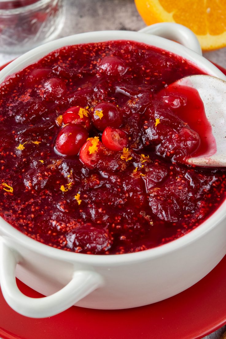 Close-up of thick homemade cranberry orange sauce with a spoon showing its chunky texture, topped with fresh orange zest and whole and halved cranberries.
