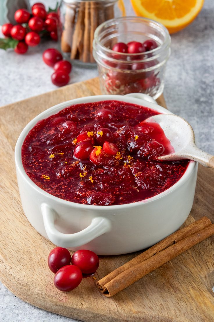 Homemade cranberry orange sauce topped with fresh orange zest in a serving bowl, with whole cranberries and cinnamon sticks in front and Christmas tree decorations, jars of cinnamon sticks, and red cranberries in the background.
