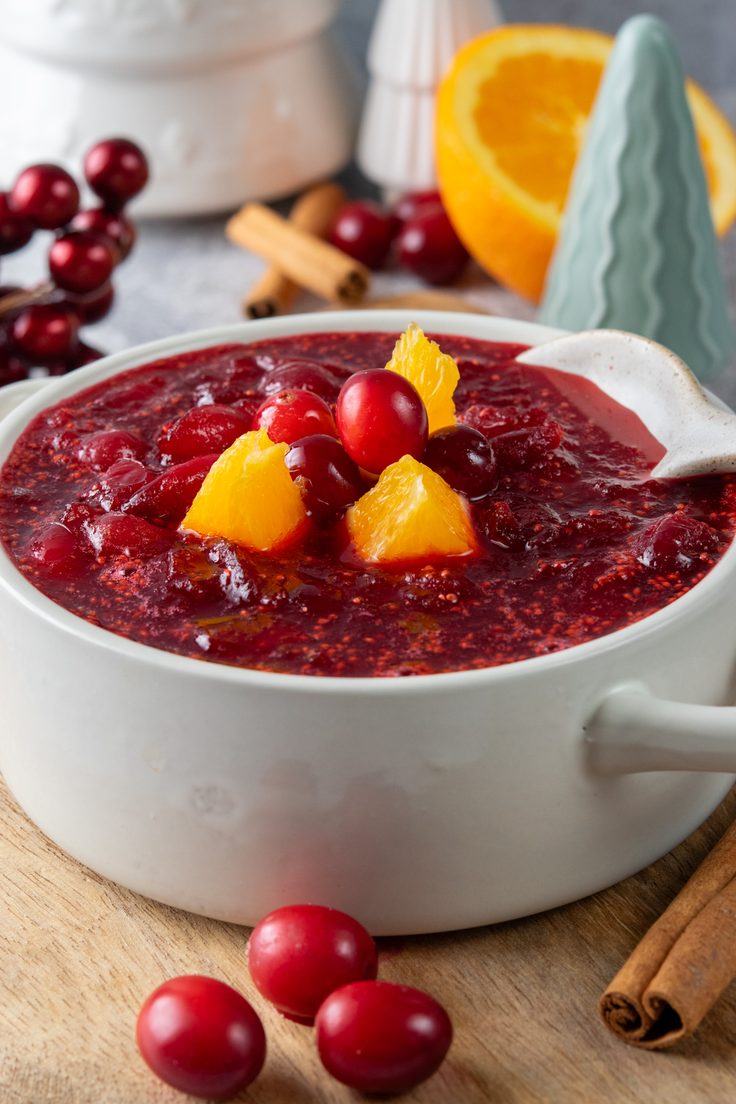 Bowl of homemade cranberry orange sauce arranged with decorative Christmas trees, ornaments, and cinnamon sticks for a festive holiday dinner.