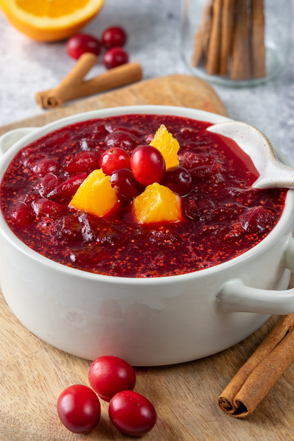 Homemade cranberry orange sauce in a small serving bowl with a spoon, garnished with fresh orange slices and whole cranberries, surrounded by cinnamon sticks, orange halves, and cranberries for a festive holiday presentation.