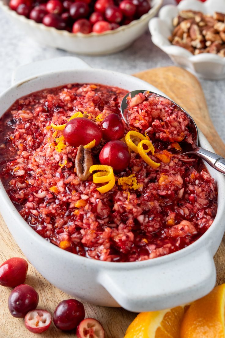 A bowl of homemade cranberry relish with a spoon scooping out a serving, surrounded by fresh cranberries, chopped pecans, and orange wedges for a colorful Thanksgiving side dish.