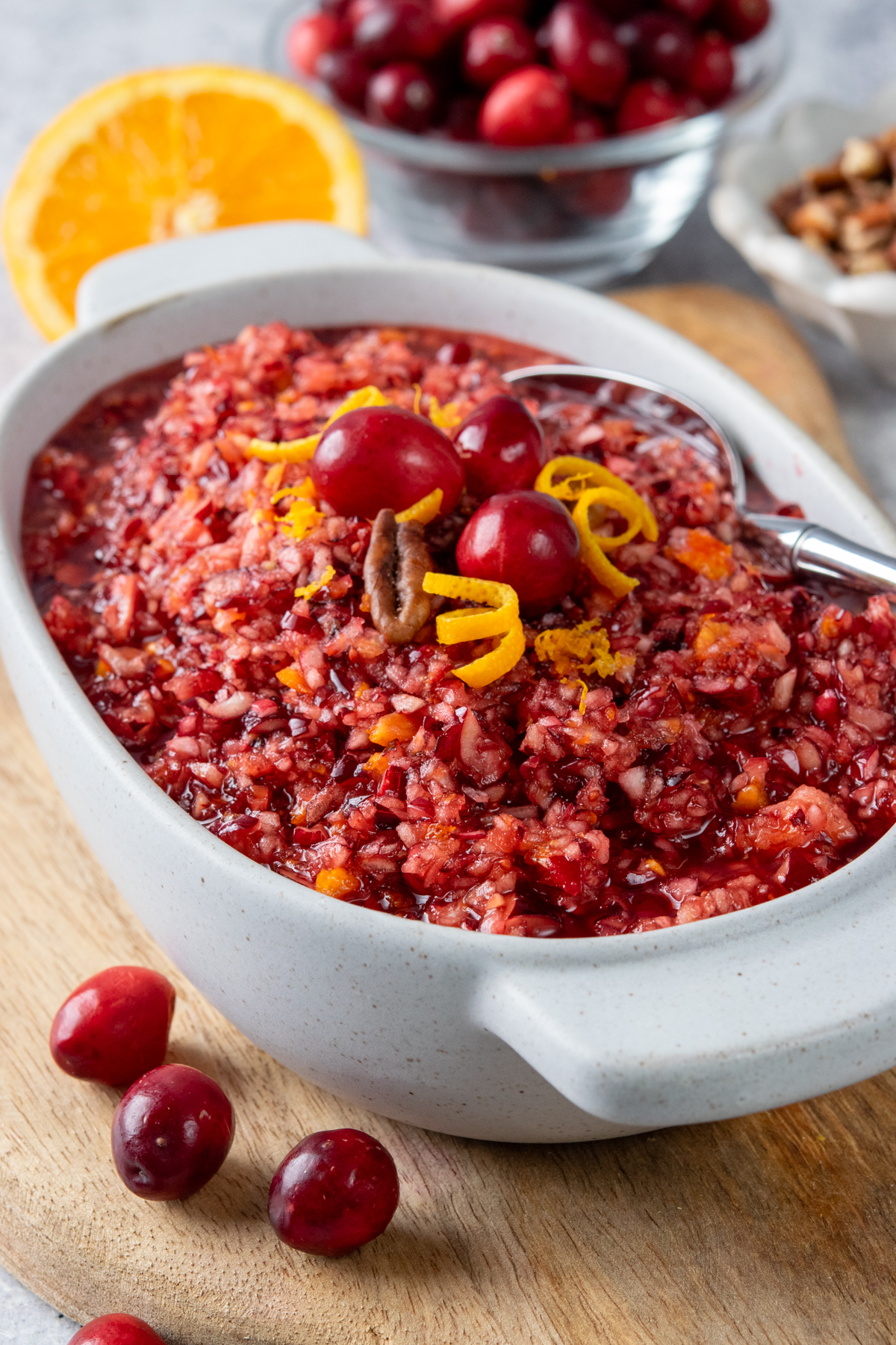 A bowl of homemade cranberry relish garnished with whole cranberries, curled orange peel, and chopped pecans, served alongside fresh cranberries, an orange, and pecans for an easy Thanksgiving condiment.